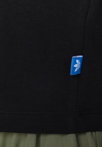 Czarna tkanina o gładkiej strukturze, z małą niebieską metką z białym logo przy szwie. Wymiarowy szczegół z złożonego ubrania pod spodem.