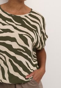 Koszulka z krótkim rękawem w zielono-kremowy wzór zebra. Tkanina ma teksturowane wykończenie, luźny krój oraz okrągły dekolt.