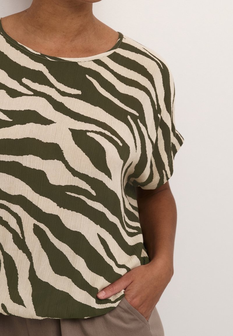 Koszulka z krótkim rękawem w zielono-kremowy wzór zebra. Tkanina ma teksturowane wykończenie, luźny krój oraz okrągły dekolt.