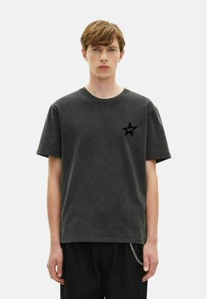 Jongeman met kort bruin haar, gekleed in een verwassen zwart T-shirt met een klein sterrenlogo op de borst en zwarte broek met een ketting.