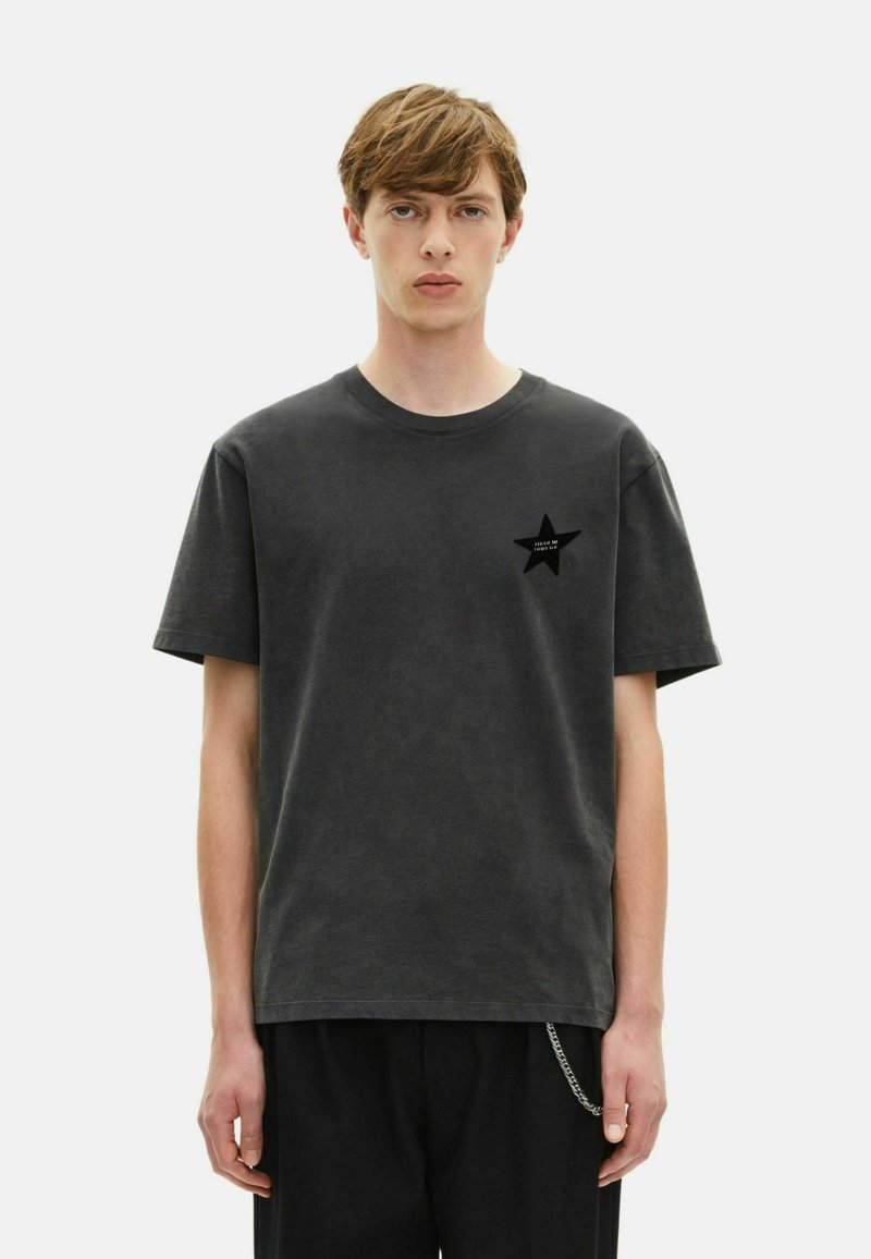 Joven con cabello corto castaño, vistiendo una camiseta negra desgastada con un pequeño logo de estrella en el pecho y pantalones negros con una cadena.