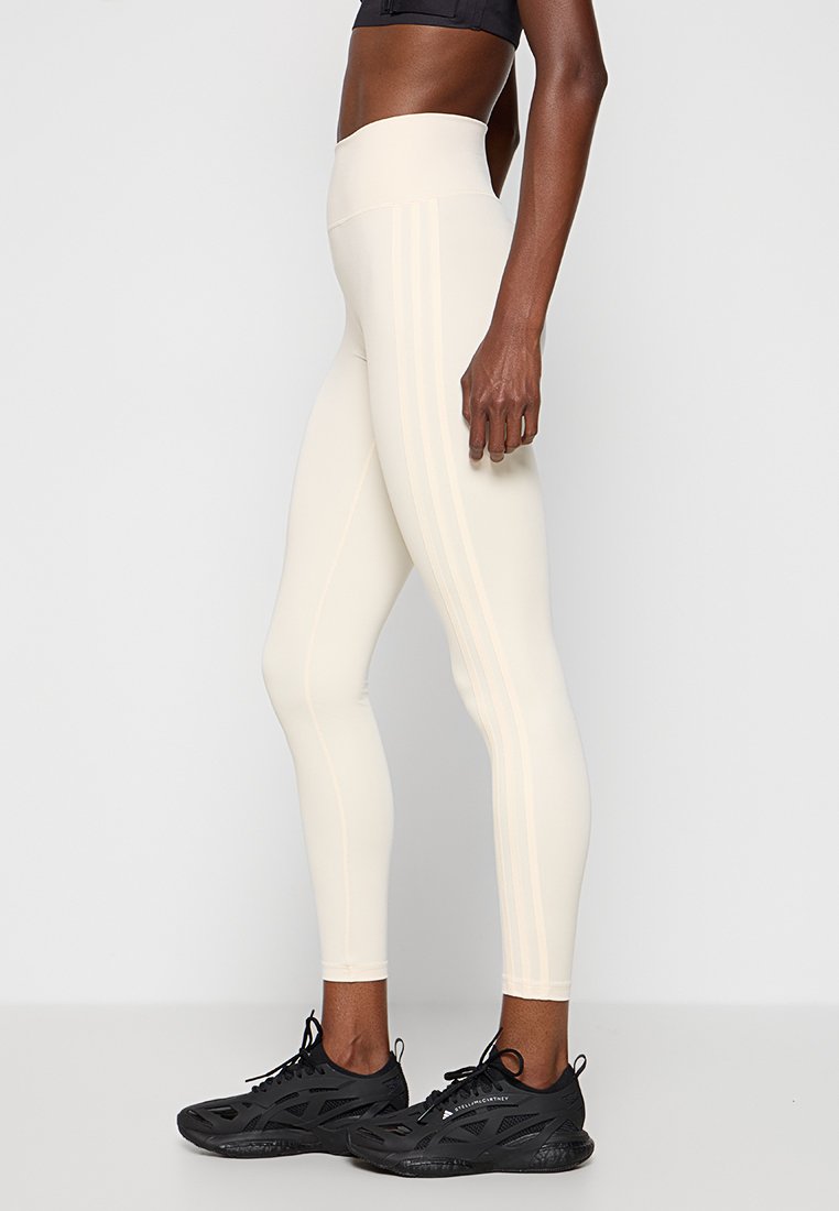 Leggings a vita alta di colore crema con una texture liscia e due righe verticali lungo ciascun lato. Abbinati a scarpe da ginnastica nere.