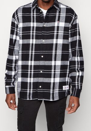 Mann trägt ein kariertes Hemd in Schwarz, Weiß und Grau mit langen Ärmeln und schwarze Cargo-Hose, steht vor einem schlichten Hintergrund.