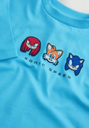 Šviesiai mėlynas medvilninis marškinėlius su siuvinėjimais, kuriuose pavaizduoti Knuckles (raudonas), Tails (oranžinis) ir Sonic (mėlynas), su tekstu "SONIC SPEED" apačioje.