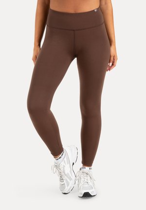 Frau trägt braune Leggings und weiße Sneakers und steht mit einem leicht über das andere gekreuzten Bein vor einem einfarbigen Hintergrund.