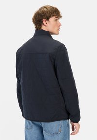 Granatowa pikowana kurtka z stójką, z wzorem w diamenty, z elastycznymi mankietami i gładkim wykończeniem. Noszona na jasnoniebieskie dżinsy.