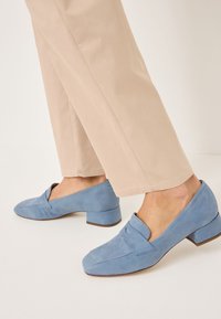 Blå mockaskor med låg klack, rund tå och ett korsat remdesign, kombinerat med ljusbeige vida byxor.