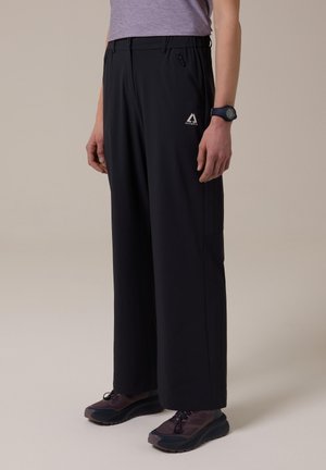 Persona con pantalones negros de pierna ancha con bolsillos con cremallera, camiseta morada, reloj deportivo negro y zapatillas de trail running moradas, de pie sobre un suelo claro.