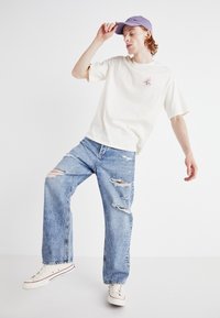 T-shirt crème avec un petit motif décoratif rose, associé à un jean délavé bleu clair. Le modèle porte une casquette violette et des baskets blanches.