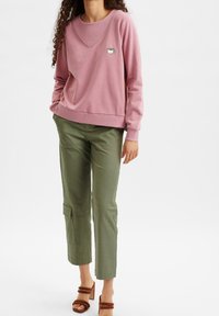 Rosa sweatshirt med V-ringning och en grafik av en kaffekopp; matchad med gröna smala byxor med en cargoficka. Bruna klacksandaler med blockklack.