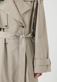 Gabardina beige, tela ligera, diseño de doble pecho, gran cuello, dos botones, cintura ceñida con cinturón, mangas largas con acentos en los puños.