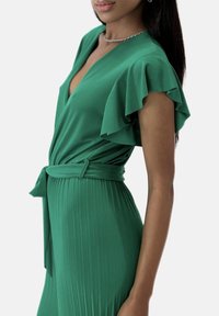 Vestido verde con mangas acampanadas, diseño cruzado y falda plisada. La textura suave y el cinturón a juego crean una silueta elegante.