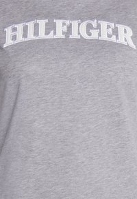 Camiseta de algodón gris con el texto bordado en blanco "HILFIGER" en el pecho. Textura suave con un ajuste estándar y escote redondo.