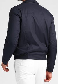 Námořnická modrá bomber bunda s hladkou, polstrovanou strukturou, žebrovanými manžetami a přiléhajícím pasem. Obsahuje nenápadné logo na rameni.