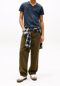 Navy V-neck T-shirt, olive green wide pants, and a blue plaid shirt tied around the waist. White and beige sneakers complete the look.