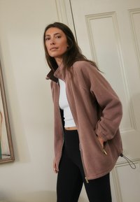 Een vrouw met lang haar draagt een lichtroze fleecejack over een witte top en zwarte leggings, terwijl ze bij een beschilderde muur staat.