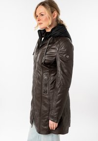 Manteau en cuir marron avec une capuche en duvet noir, fermeture éclair, poches latérales et design ajusté. Présente des détails de couture et une finition texturée.
