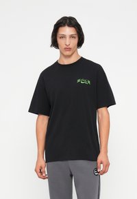 Jauns vīrietis ar plecu garuma matiem, tērpts melnā Nike SB kreklā ar zaļu logotipu un pelēkās Nike sporta biksēs uz vienkārša baltā fona.