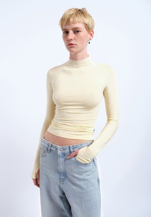 Long-sleeve, mock neck top in pale yellow, made from a smooth, fitted fabric. Paired with light blue high-waisted jeans with a relaxed fit.