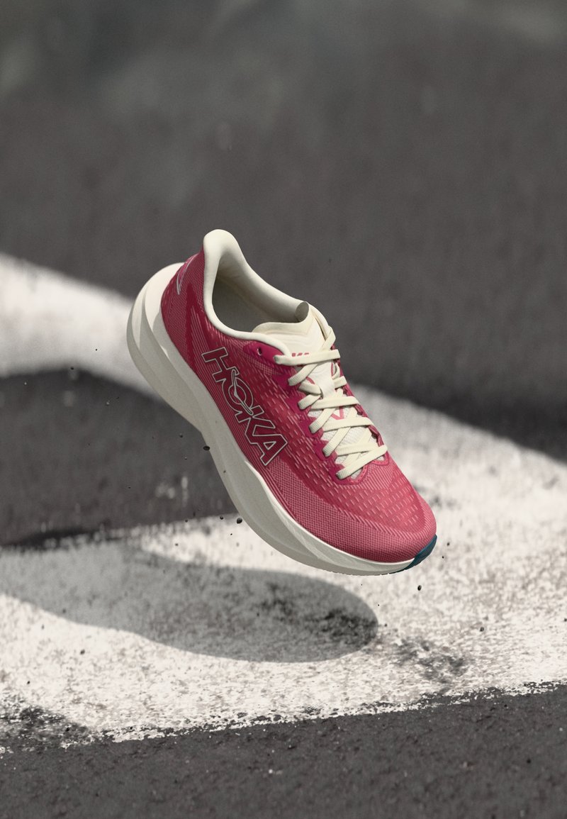 Rødt og hvitt HOKA-løpesko henger over asfalt med hvite veimerkninger, små gruspartikler flyr rundt sålen.