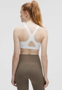 Balta sportinė liemenėlė su lenktu pečių dizainu, turinti raktų skylutės išpjovimą ir elastingą juostą. Derinama su aukštu liemeniu rudomis legging'ais.