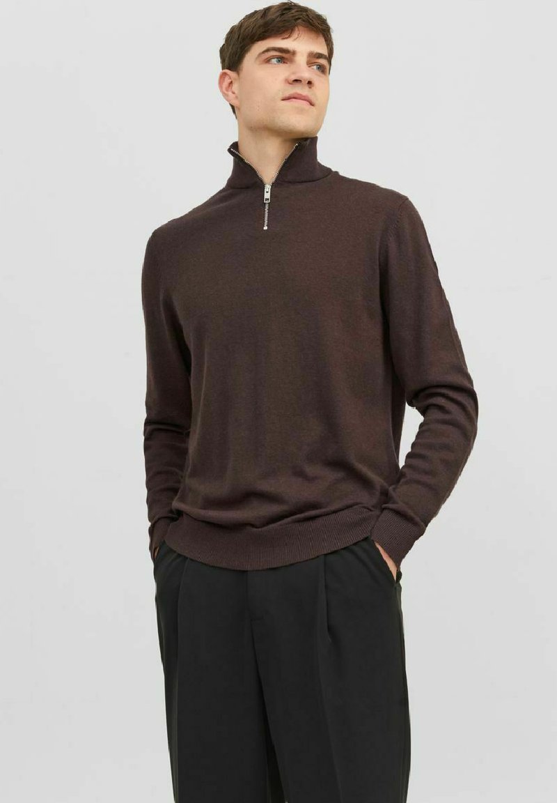 Pull en tricot marron avec un col haut, doté d'une fermeture éclair. Poignets et ourlet côtelés. Porté avec un pantalon noir ajusté.