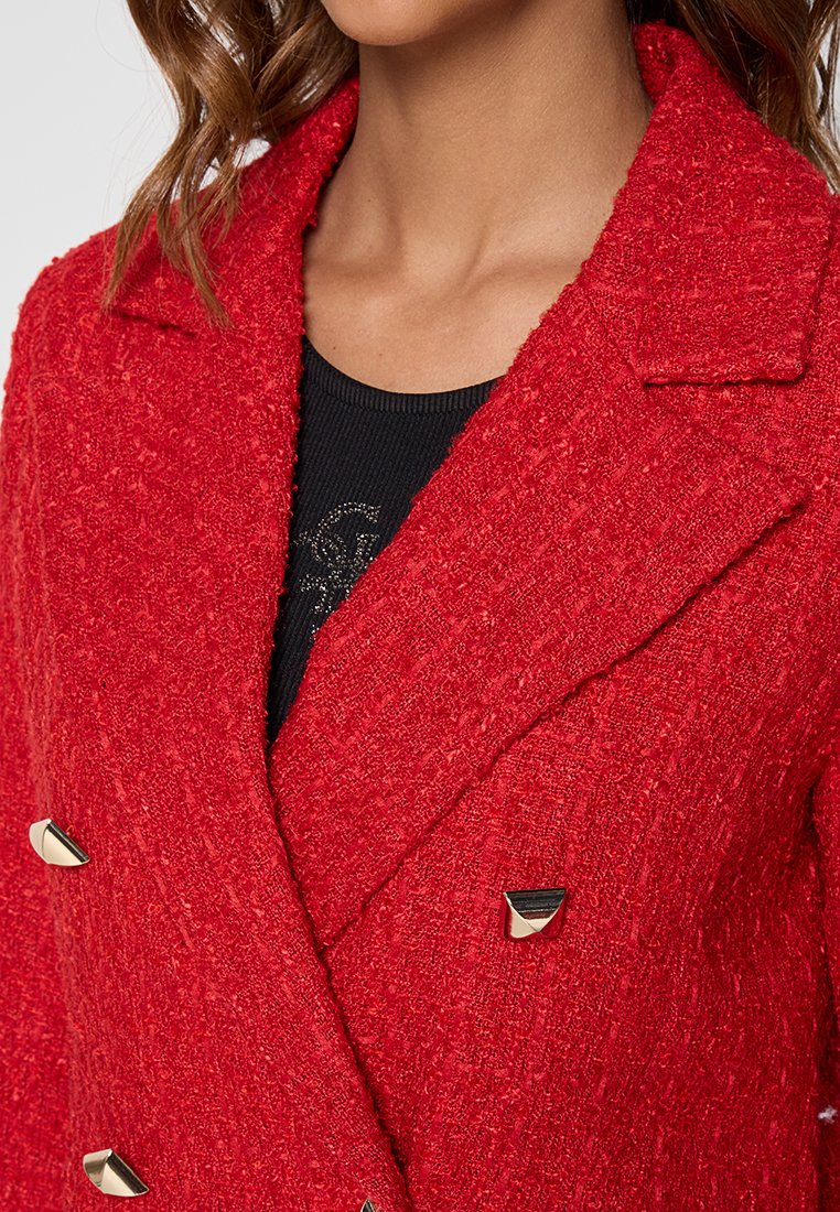 Cappotto double-breasted rosso con texture, colletto pronunciato e accenti in hardware dorato. Indossato sopra una maglietta nera con dettagli sottili.