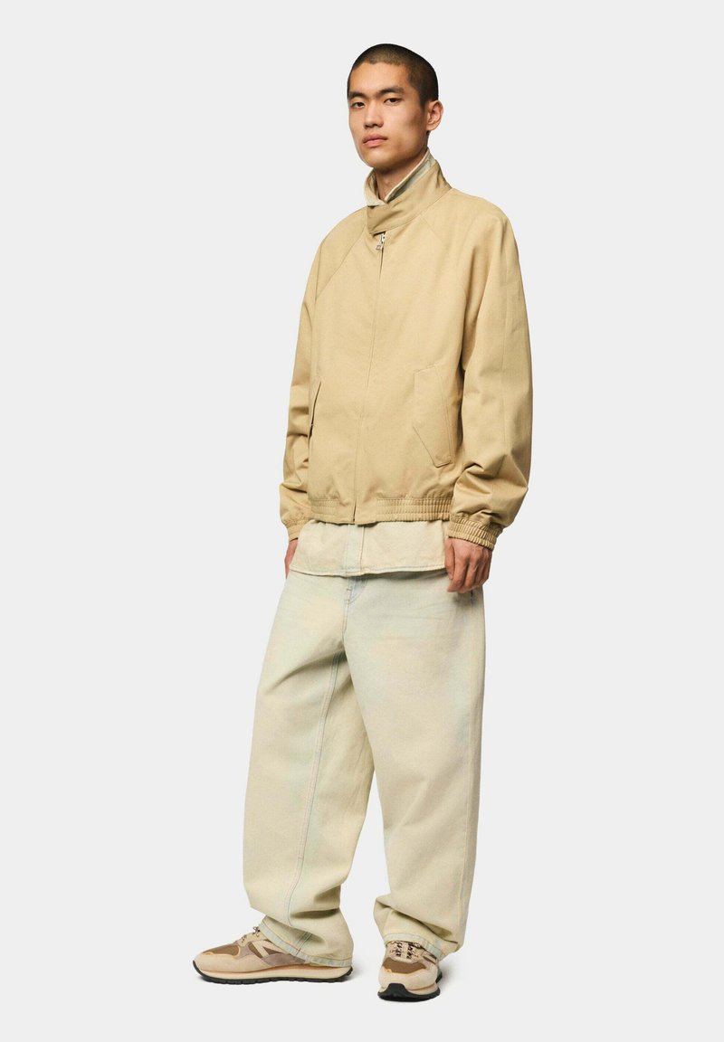 Veste bomber beige avec un col montant, portée sur une chemise de couleur claire et un pantalon en denim pâle, ample. Baskets marron et beige.