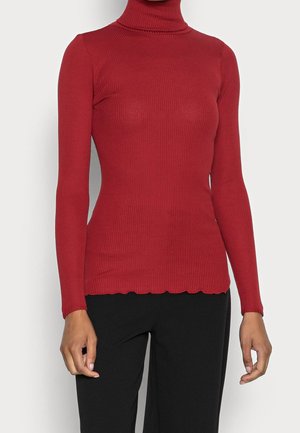 Mujer vistiendo un suéter rojo ajustado de cuello alto y tejido acanalado, junto con pantalones negros, mostrada desde el cuello hasta las caderas sobre un fondo liso.