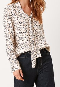 Blusa a maniche lunghe in tessuto leggero, con stampa geometrica multicolore, fiocco al collo e vestibilità rilassata abbinata a jeans scuri.