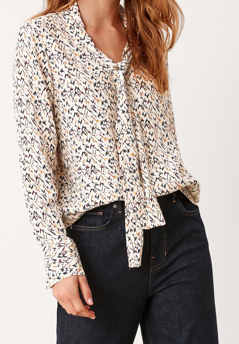 Blusa a maniche lunghe in tessuto leggero, con stampa geometrica multicolore, fiocco al collo e vestibilità rilassata abbinata a jeans scuri.
