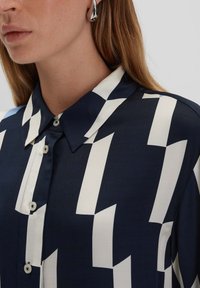 Camicia di seta blu navy con un motivo geometrico bianco, chiusura con bottoni e colletto classico. Texture liscia con una finitura lucida.