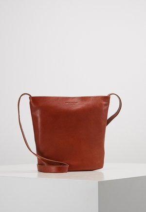 Brązowa torebka wiaderkowa ze skóry z długim paskiem, gładką fakturą i subtelnie wytłoczonym nazwą marki na przedzie, prezentowana na białym piedestale.