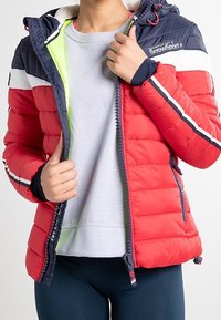 Rød fyldig jakke med marineblå detaljer, quiltet design, lynlås lukning og hætte. Har en lys grøn foring og hvide striber på ærmerne.