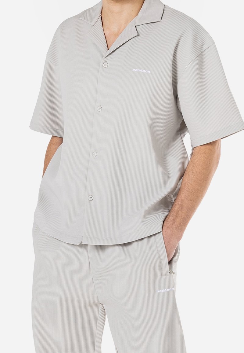Homme portant une chemise côtelée gris clair à manches courtes boutonnée et un pantalon côtelé assorti, les mains dans les poches.