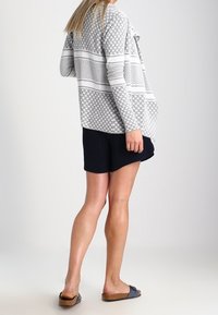 Šedý pletený kardigan s geometrickými vzory, volně přehnutý vpředu, dlouhé rukávy, nošený přes černé šortky, doplněný o nazouvací sandály.