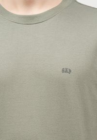 Κοντινή λήψη ενός μπλουζιού σε χρώμα ανοιχτό ελιάς με στρογγυλή λαιμόκοψη και ένα μικρό, μαύρο λογότυπο «GAP» στο αριστερό μέρος του στήθους.