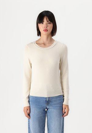 Hellbeiger Pullover aus weichem Material mit rundem Ausschnitt, der perlenartige Akzente und lange Ärmel aufweist, kombiniert mit blauen Jeans.
