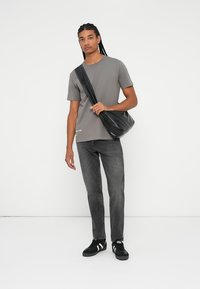 Camiseta gris de manga corta, bolso cruzado negro, jeans gris oscuro y zapatillas negras con rayas blancas, de pie contra un fondo liso.