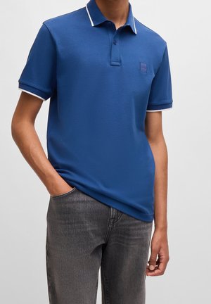 Mężczyzna w niebieskiej koszulce polo z krótkim rękawem z białym wykończeniem na kołnierzyku i rękawach, w połączeniu z wyblakłymi czarnymi dżinsami, z ręką w kieszeni.