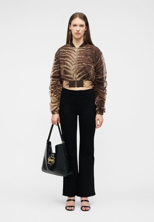 Vrouw draagt een cropped bomberjack met bruin patroon, zwarte broek, zwarte sandalen met bandjes, houdt een zwarte handtas met gouden sluiting vast, staat tegen een witte achtergrond.