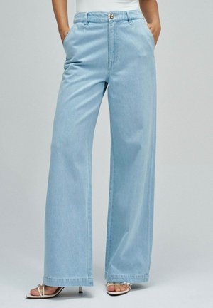 Frau trägt hellblaue Jeans mit weitem Bein und vorderen Taschen, kombiniert mit weißen, geschnürten Sandaletten mit Absatz, steht vor schlichtem Hintergrund.