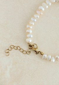 Bracelet en perles comprenant des perles blanches irrégulières et un fermoir en or avec une extension en chaîne pour un ajustement de la longueur. Surface texturée, formes arrondies.