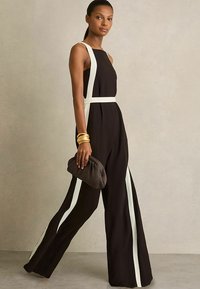 Zwart jumpsuit met witte zijkanten, mouwloos ontwerp, wijde pasvorm, gladde textuur. Model houdt een gestructureerde bruine clutch tas vast.