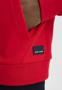 Felpa rossa con polsini e orlo a costine. Presenta una targhetta blu navy con il logo "JOOP! JEANS" cucita sul lato. Tessuto in misto cotone.