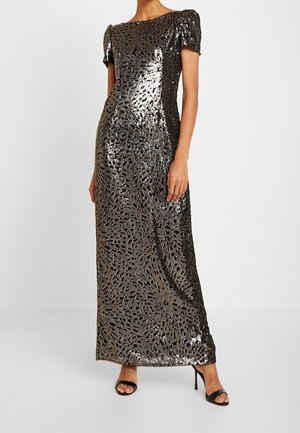 Femme portant une robe longue noire et argentée à manches courtes ornée de sequins, ainsi que des talons hauts noirs ouverts, debout devant un fond blanc.