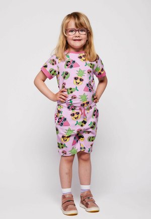 Młoda dziewczynka w okularach stoi z rękami na biodrach, ubrana w różowe krótkie spodenki i koszulkę z nadrukiem uśmiechniętych owocowych postaci w okularach przeciwsłonecznych.