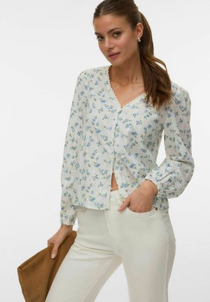 Kobieta z długimi brązowymi włosami związanymi w kucyk nosi białą bluzkę z niebieskim kwiatowym wzorem i białe spodnie, trzymając w jednej ręce brązową kopertówkę.