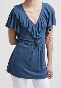 Niebieska bluzka z krótkim rękawem, z głębokim dekoltem w kształcie litery V i falbaniastymi detalami wzdłuż dekoltu oraz rękawów. Miękka tkanina z marszczonym pasem.