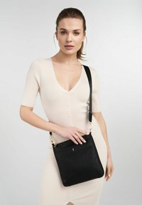 Kobieta w kremowej sukience z prążkowanego materiału, zapinanej na guziki, trzyma czarną skórzaną torebkę crossbody ze złotymi akcentami i szerokim paskiem.